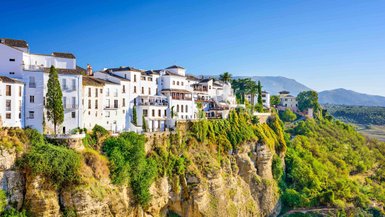 Die weißen Gebäude von Ronda thronen majestätisch auf einer Klippe, umgeben von üppigem Grün und strahlend blauem Himmel.