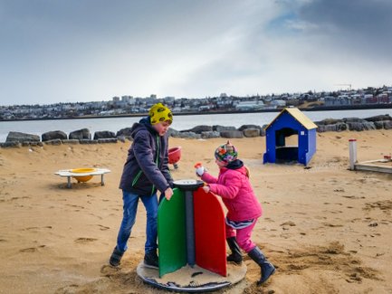 Zwei Kinder spielen auf einem Sandspielplatz, während im Hintergrund eine Stadtlandschaft und ein ruhiger Wasserbereich sichtbar sind.