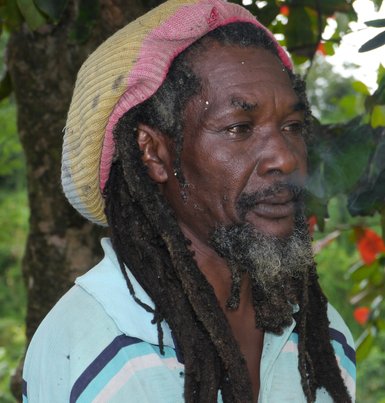 Ein Mann mit langen, lockigen Haaren und einem bunten Hut steht nachdenklich vor einer grünen, natürlichen Kulisse.