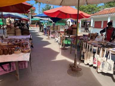 Bunte Marktstände mit handgefertigten Waren, darunter Hüte, Holzspielzeug und Taschen, unter sonnigen Schirmen.