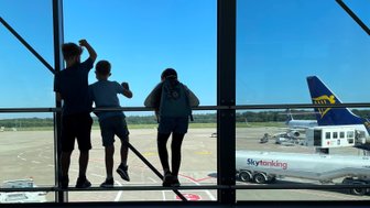 Drei Kinder stehen am Fenster eines Flughafens und beobachten das Geschehen auf dem Rollfeld bei strahlend blauem Himmel.