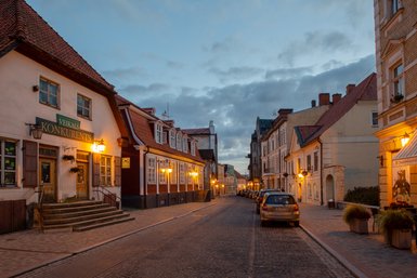 Eine malerische Straße in Cesis, beleuchtet von warmen Laternen, umgeben von historischen Gebäuden und einem bewölkten Himmel.