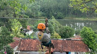 Ein Abenteurer schwingt an einer Zipline über ein malerisches Tal mit üppigem Grün und einem ruhigen See im Hintergrund.