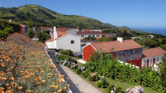 Bunte Blumen schmücken den Weg zu charmanten Häusern, umgeben von sanften Hügeln und dem blauen Meer im Hintergrund.