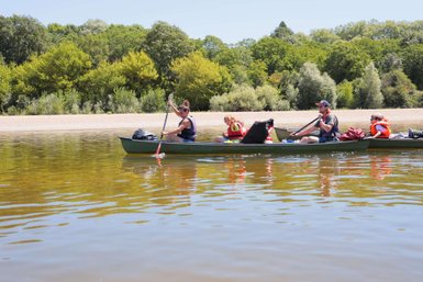 Eine Gruppe von vier Personen paddelt in einem Kanu auf einem ruhigen Fluss, umgeben von üppigem Grün und klarem Himmel.
