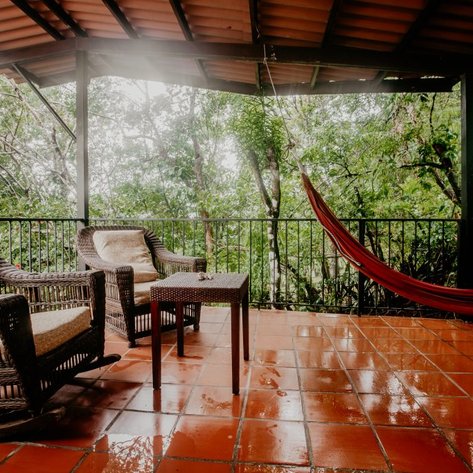 Eine gemütliche Terrasse mit zwei Korbsesseln, einem Tisch und einer Hängematte, umgeben von üppigem Grün.