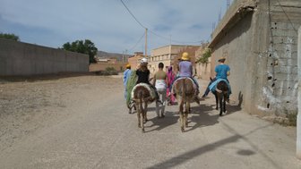 Eine Gruppe von Menschen reitet auf Eseln durch eine staubige Straße in einem marokkanischen Dorf.
