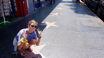 Eine junge Frau kniet auf dem Hollywood Boulevard und zeigt das Peace-Zeichen, umgeben von Sternen auf dem Gehweg.