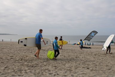 Ein Erwachsener und ein Kind tragen Surfbretter am Strand, während andere Surfer im Hintergrund aktiv sind.