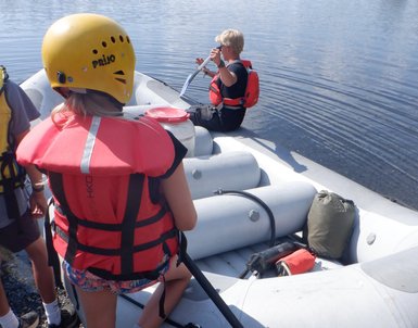 Eine Gruppe von Personen bereitet sich auf eine Raftingtour auf einem ruhigen Gewässer mit einem Schlauchboot vor.