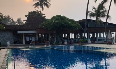 Ein ruhiger Pool spiegelt die Palmen und die untergehende Sonne wider, während der Himmel in sanften Farben leuchtet.