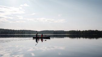 Zwei Personen paddeln in einem Kanu auf einem ruhigen See, umgeben von dichten Wäldern und einem klaren Himmel.