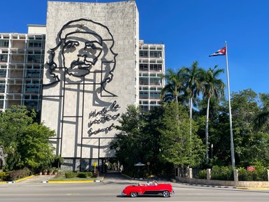 Ein rotes Cabrio fährt vorbei an einem großen Wandbild von Che Guevara, umgeben von Palmen und einem blauen Himmel.