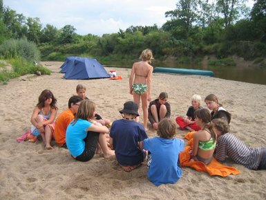 Eine Gruppe von Kindern sitzt im Sand am Flussufer, während sie an einer Campbesprechung teilnehmen und sich unterhalten.