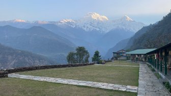 Eine weite Aussicht auf schneebedeckte Berge, die über eine grüne Wiese und ein Steingebäude im Vordergrund ragt.