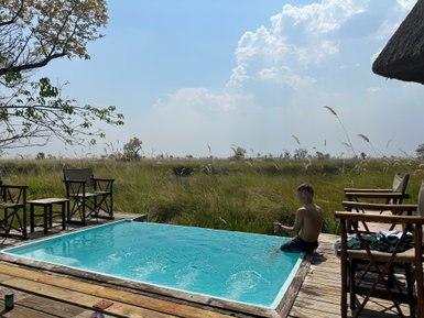 Ein Junge sitzt am Rand eines Pools und spielt mit Wasser, umgeben von hohen Gräsern und einem klaren Himmel.