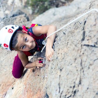 Ein junges Mädchen klettert an einer Felswand, konzentriert und mit einem Helm, während sie sich an einem Seil festhält.