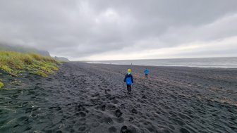 Zwei Kinder in blauen Jacken laufen über einen schwarzen Sandstrand, umgeben von grauen Wolken und sanften Hügeln.