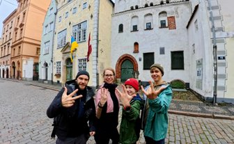 Vier Personen posieren fröhlich auf einer gepflasterten Straße in Riga, umgeben von historischen Gebäuden.