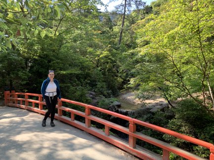 Eine Frau steht an einer roten Brücke, umgeben von üppigem Grün und Bäumen, während ein kleiner Wasserlauf im Hintergrund fließt.