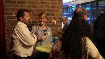 Eine Frau mit einem Drink lächelt, während sie mit einem Mann an einem Tisch in einer geselligen Umgebung spricht.