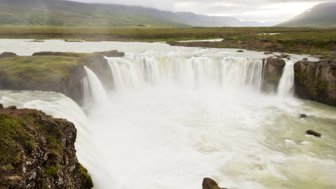 Der beeindruckende Godafoss-Wasserfall stürzt in einen breiten Fluss, umgeben von üppigem, grünem Gras und sanften Hügeln.