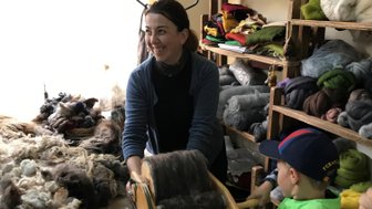 Eine fröhliche Frau und ein Junge arbeiten gemeinsam an einem Wollprojekt in einem hellen, gemütlichen Raum voller Materialien.