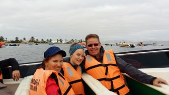 Drei Personen in orangefarbenen Schwimmwesten lächeln auf einem Boot, umgeben von Wasser und kleinen Booten im Hintergrund.