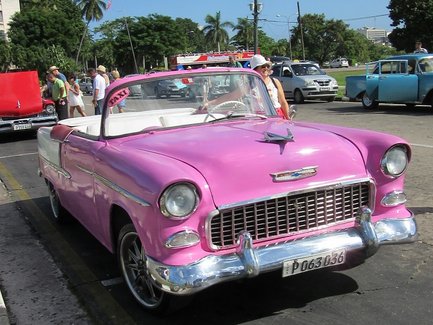 Ein leuchtend pinkfarbener Oldtimer steht auf einer Straße, umgeben von Palmen und Menschen, die die Szene genießen.