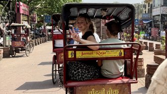 Eine Frau mit blonden Haaren lächelt und zeigt ein Peace-Zeichen, während sie in einer Fahrradrikscha sitzt.