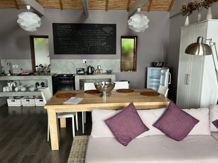 Eine moderne Küche mit einem großen Holztisch, weißen Stühlen und lila Kissen auf einer gemütlichen Couch im Hintergrund.