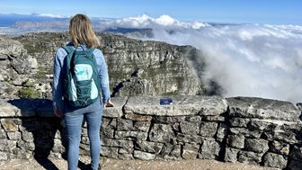 Eine Person mit einem Rucksack steht an einer Steinmauer und blickt auf eine beeindruckende Berglandschaft und Wolken.