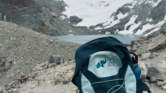 Ein Rucksack steht auf einem großen Stein, umgeben von schneebedeckten Bergen und einem klaren See im Hintergrund.