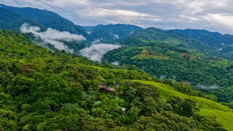 Dichte grüne Wälder umgeben eine kleine Lodge, die idyllisch zwischen sanften Hügeln und Bergen liegt.