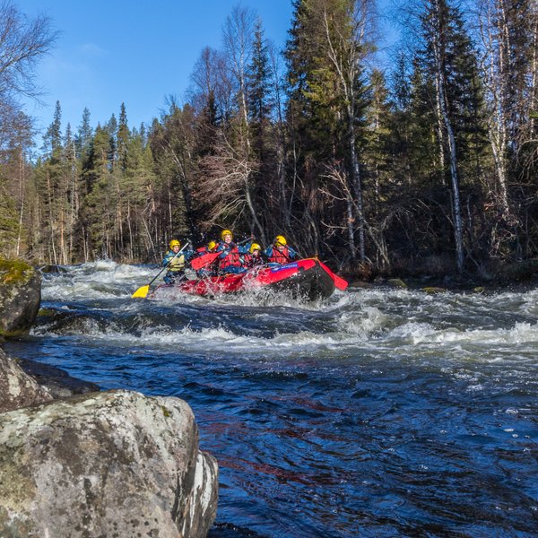 Eine Gruppe von vier Personen paddelt in einem roten Raft durch einen reißenden Fluss, umgeben von grünen Bäumen.