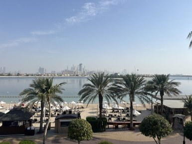 Ein ruhiger Strand mit Palmen und Liegen, während die Skyline von Dubai im Hintergrund sichtbar ist.