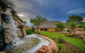 Eine malerische Lodge mit einem Wasserfall und üppigem Grün, umgeben von tropischen Pflanzen und einem bewölkten Himmel.