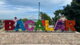 Drei Personen posieren fröhlich vor einem bunten 'Bacalar'-Schild, umgeben von einer malerischen Landschaft und blauem Himmel.