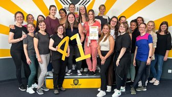 Eine Gruppe von Frauen steht fröhlich auf einer Bühne, umgeben von gelben Strahlen an der Wand, und hält ein Zertifikat.