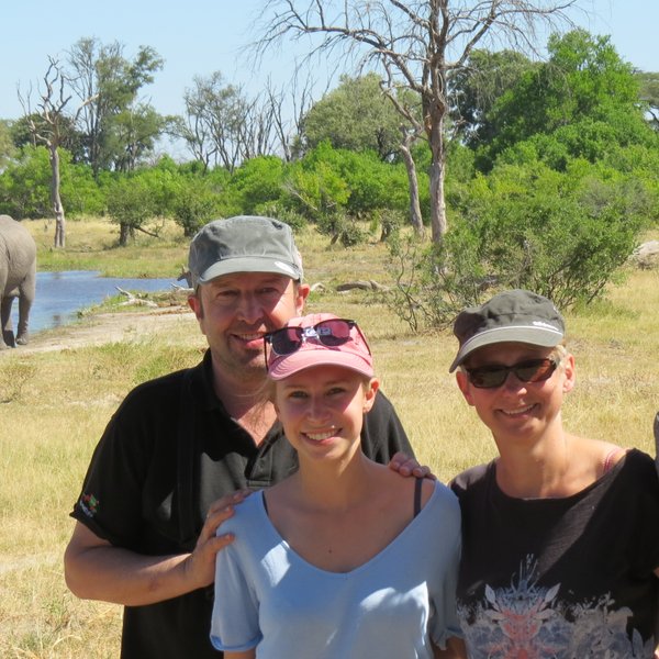 Eine Familie steht lächelnd vor einem Elefanten, der im Hintergrund an einem Fluss steht, umgeben von grüner Natur.