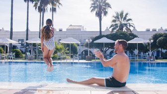 Ein Kind springt fröhlich ins Wasser, während ein Mann am Beckenrand sitzt und lächelt, umgeben von Palmen und Sonnenschirmen.