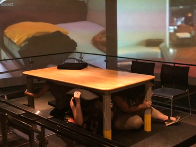 Unter einem Tisch kauern mehrere Personen, während im Hintergrund eine Erdbebensimulation auf einer Leinwand gezeigt wird.