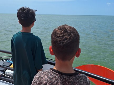 Zwei Jungen stehen auf einem Boot und blicken auf das ruhige Wasser des Meeres unter einem klaren blauen Himmel.