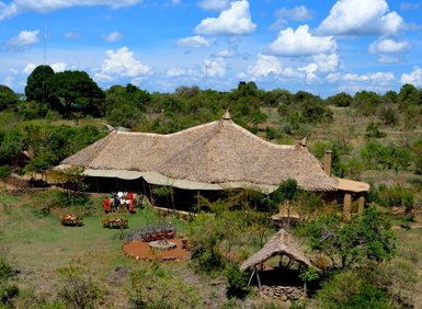 Eine luftige Ansicht eines traditionellen, strohgedeckten Camps inmitten einer grünen, bewaldeten Landschaft unter blauem Himmel.