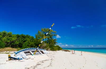 Ein verlassenes Boot liegt auf dem weißen Sandstrand, während eine Familie am Ufer entlang spaziert und die Sonne genießt.