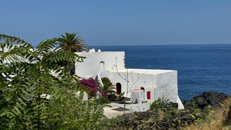 Ein Blick auf eine weiße Villa mit roten Akzenten, umgeben von üppigem Grün und dem blauen Meer im Hintergrund.