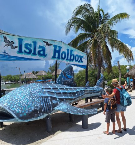 Ein großes, buntes Modell eines Walschwanzes steht unter dem Schild 'Isla Holbox', während eine Familie es bewundert.