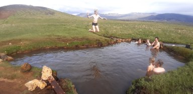 Ein Junge springt fröhlich in eine heiße Quelle, während andere im Wasser entspannen und die Natur genießen.