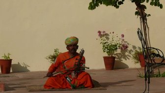 Ein indischer Straßenmusiker sitzt im Freien, trägt ein leuchtendes orangefarbenes Gewand und spielt ein traditionelles Instrument.