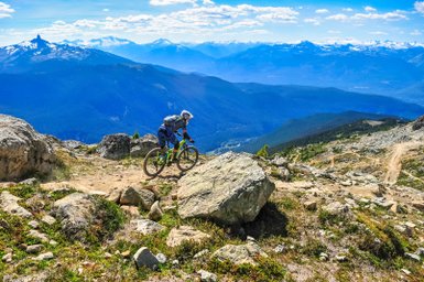 Ein Mountainbiker fährt über einen steinigen Pfad mit beeindruckender Berglandschaft im Hintergrund.